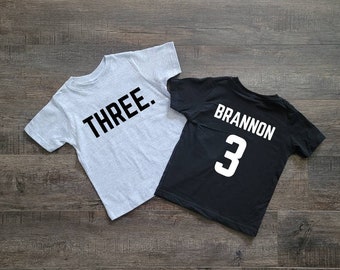 May include: Two gray and black t-shirts with the words "THREE." and "BRANNON 3" printed on them.