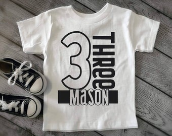 May include: White t-shirt with black text and a large black number 3. The text reads "Three Mason".