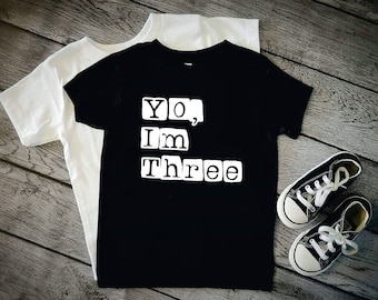 May include: Black t-shirt with white text that reads "Yo, I'm Three" in a blocky font. The shirt is on a wooden surface with a pair of black sneakers.