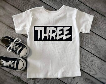 May include: White t-shirt with black text that says "THREE" in a bold, graffiti-style font.