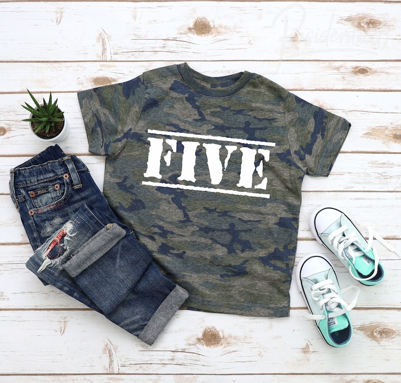 May include: A camouflage print t-shirt with the word "FIVE" printed in white on the front. The shirt is laid out on a white wooden surface with a pair of blue jeans and two pairs of light blue sneakers.