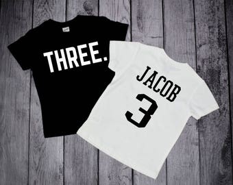 May include: Two black and white t-shirts. One shirt says "THREE." The other shirt says "JACOB" and has the number "3" on it.