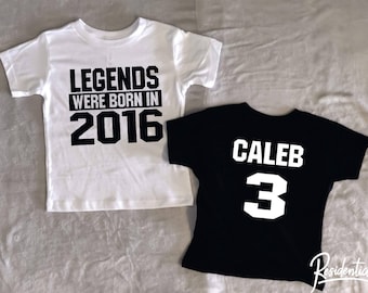 May include: Two white t-shirts with black text. One shirt says "Legends were born in 2016". The other shirt is black with white text that says "Caleb 3".