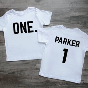 May include: Two white short-sleeved t-shirts. The first shirt features the black text "ONE." centered on the chest. The second shirt features the black text "PARKER" above the number "1" in a bold, sans-serif font on the chest area.