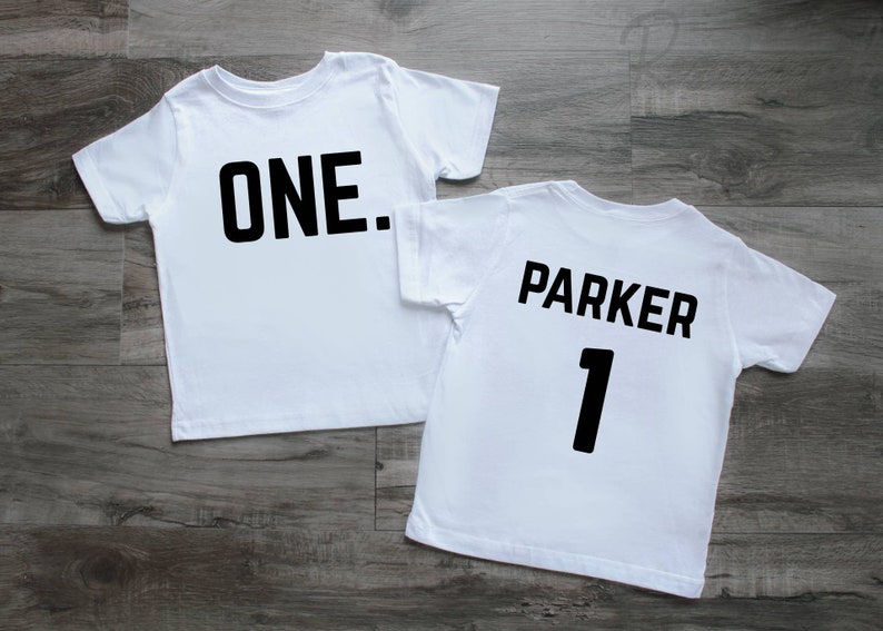May include: Two white short-sleeved t-shirts. The first shirt features the black text "ONE." centered on the chest. The second shirt features the black text "PARKER" above the number "1" in a bold, sans-serif font on the chest area.