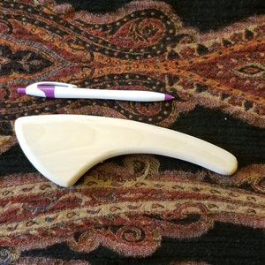 May include: A white, curved, wooden tool for massage therapy. The tool is about 12 inches long and 3 inches wide at its widest point. It is lying on a patterned fabric.