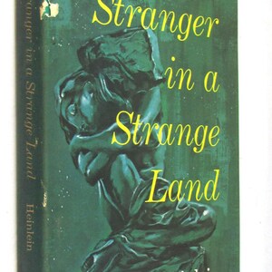 Peut inclure: Un livre de poche vintage intitulé "Stranger in a Strange Land" de Robert A. Heinlein. La couverture est vert foncé avec des lettres jaunes et une figure sculptée. La tranche du livre affiche également le titre et l'auteur.