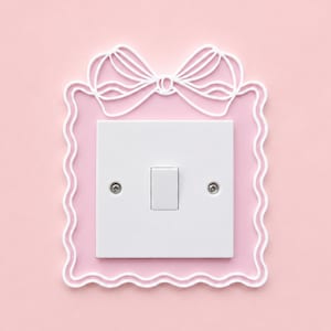 May include: A white light switch plate cover featuring a decorative pink square frame with a scalloped edge and a white bow design at the top. The switch plate is centred within the frame and includes two silver screws on the left and right sides.