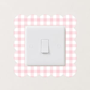 May include: A white light switch plate featuring a central toggle switch and two circular screw covers, framed by a square decorative border with a scalloped edge and a light pink and white gingham checkered pattern on the border surface.