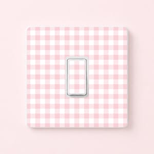 May include: A light pink and white gingham pattern light switch cover with a single toggle switch. The cover is square with rounded corners and a white toggle switch. The background is a solid light pink.