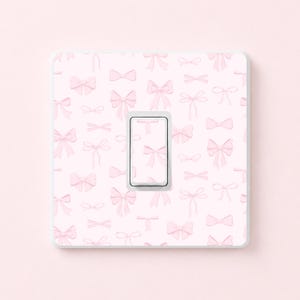 May include: A light pink light switch cover featuring a pattern of pink bows. The switch has a white frame and a pink bow design. The background is a solid light pink.