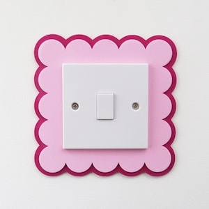 May include: A white light switch plate mounted on a wall, framed by a decorative square border with a scalloped edge. The border consists of two layers: a larger dark magenta base layer and a smaller light pink top layer.