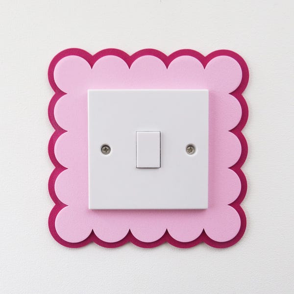 Puede incluir: Un interruptor de luz blanco montado en una pared, enmarcado por un borde cuadrado decorativo con un acabado festoneado. El borde consta de dos capas: una capa base de color magenta oscuro más grande y una capa superior rosa claro.