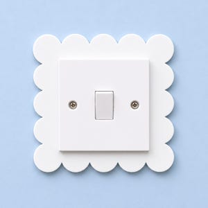 Könnte beinhalten: Eine weiße Lichtschalterabdeckung mit einem quadratischen Mittelteil, einem einzelnen Kippschalter und zwei sichtbaren Metallschrauben, umgeben von einem dekorativen Rand mit Wellenmuster. Das gesamte Element besteht aus weißem Material.