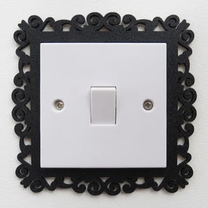 May include: A white light switch with a grey toggle, set within a black, ornate, square frame. The frame features a decorative, swirling design around the edges. The switch plate is secured with two silver screws.