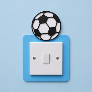 May include: A white light switch with a rectangular toggle, set within a light blue square, with a black and white football design above. The background is a solid light blue.