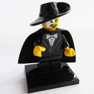 May include: A Lego figure of a character in a black tuxedo, cape, and hat, with a white mask. The figure has yellow hands and a yellow head. The figure is standing on a black base.