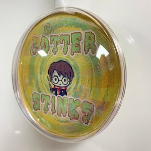 May include: A round, clear plastic ornament with a yellow and green pixelated design. The design features a cartoon character with glasses and the text "Potter Stinks".