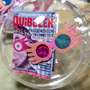Peut inclure: Une boule de Noël en verre transparente contenant des lunettes à thème Harry Potter en paillettes roses et dorées. La boule contient également un petit article de journal du journal fictif "The Quibbler" avec le titre "Pandémonium au ministère - Quel remue-ménage !"