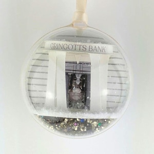 May include: A clear glass ornament with a white background and a paper cutout of Gringotts Bank, a fictional bank from the Harry Potter series. The ornament is filled with gold glitter and colorful sequins.