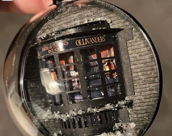May include: A glass ornament with a snowy scene depicting Ollivanders Wand Shop from the Harry Potter series. The shop's sign reads "Ollivanders Makers of Fine Wands Since 382 B.C."
