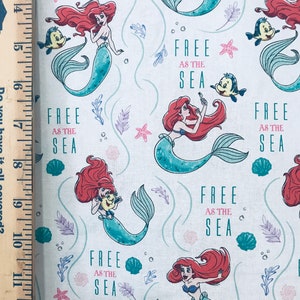 May include: A white fabric featuring a repeating pattern of a red-haired mermaid and a small yellow fish. The text "FREE AS THE SEA" is printed in teal and pink. A wooden ruler is visible on the left side, showing measurements in inches.
