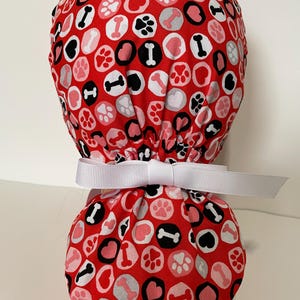 May include: A red scrub cap featuring a repeating pattern of white, pink, and black circles containing paw prints, hearts, and bones. The cap has a gathered back section secured with a plain white ribbon tied in a bow at the center.