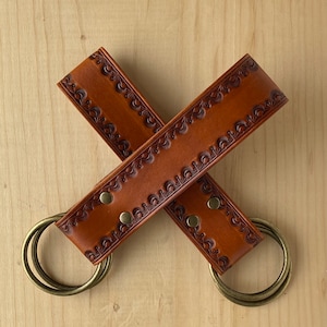 May include: Two brown leather keychains with intricate floral designs and brass rings. The keychains are crossed over each other.