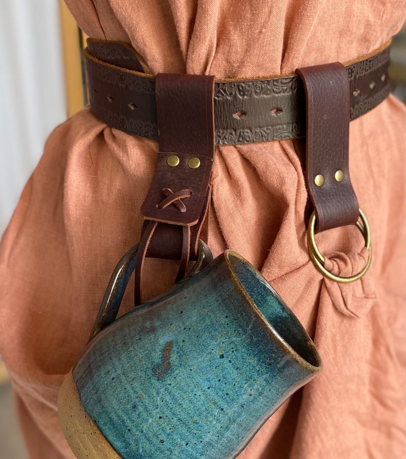 May include: A brown leather belt with a decorative design and two loops. A blue and green ceramic mug is hanging from one of the loops.