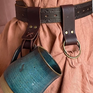 May include: A brown leather belt with a decorative design and a metal ring. A blue ceramic mug is attached to the belt with a leather strap.