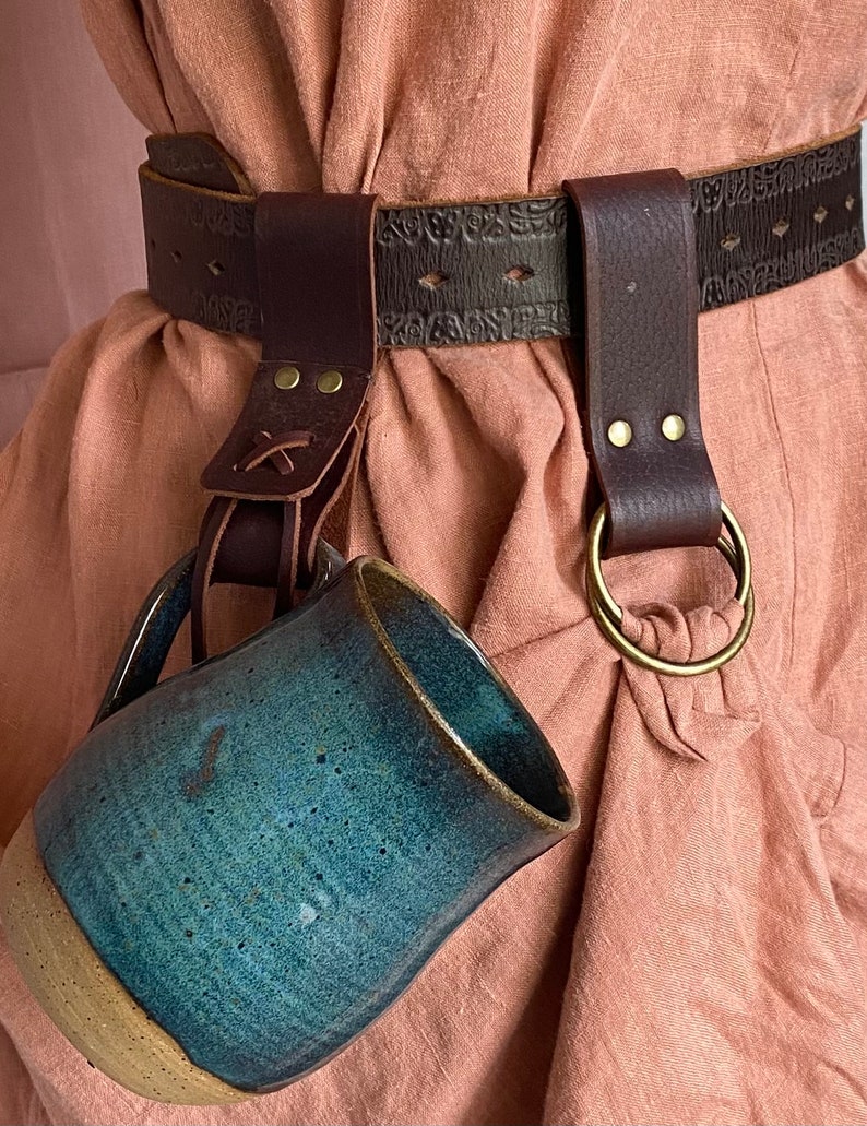 May include: A brown leather belt with a decorative design and a metal ring. A blue ceramic mug is attached to the belt with a leather strap.