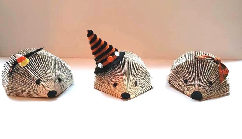 Pode incluir: Tr&ecirc;s ornamentos de ouri&ccedil;o feitos com p&aacute;ginas de livros decorados para o Halloween. Um tem um chap&eacute;u de bruxa, outro tem um milho doce e outro tem um la&ccedil;o.