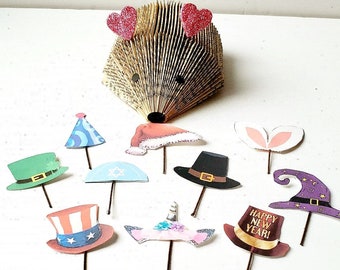 May include: A whimsical hedgehog sculpture made from book pages, adorned with glittery red heart accents. Assorted paper cutouts of hats and headwear on sticks, including a party hat, Santa hat, and a witch's hat. The website address www.treecycledcrafts.com is visible.