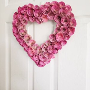 May include: A pink paper heart wreath made of rolled paper flowers. The wreath is hanging on a white door.