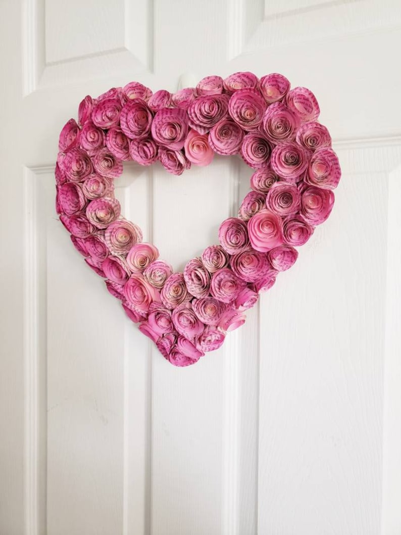 May include: A pink paper heart wreath made of rolled paper flowers. The wreath is hanging on a white door.