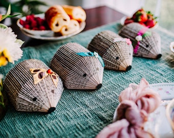 May include: Four book page hedgehog ornaments with different colored ribbons and embellishments. The hedgehogs are sitting on a teal textured surface.