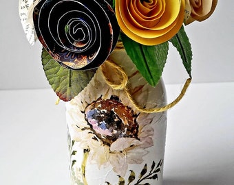 May include: A bouquet of paper flowers in shades of yellow, black, and brown, arranged in a white ceramic vase with a sunflower design and a blue base. The vase is decorated with a brown twine bow.