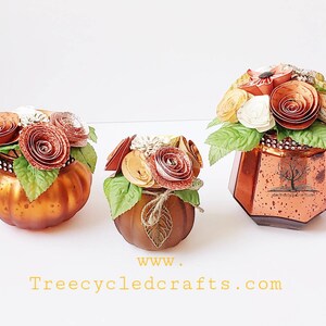 May include: Three glass pumpkins decorated with paper flowers in shades of orange, brown, and white. The pumpkins are all different sizes and have a rustic, vintage look. The largest pumpkin has a label that reads "Treecycled Crafts.com".