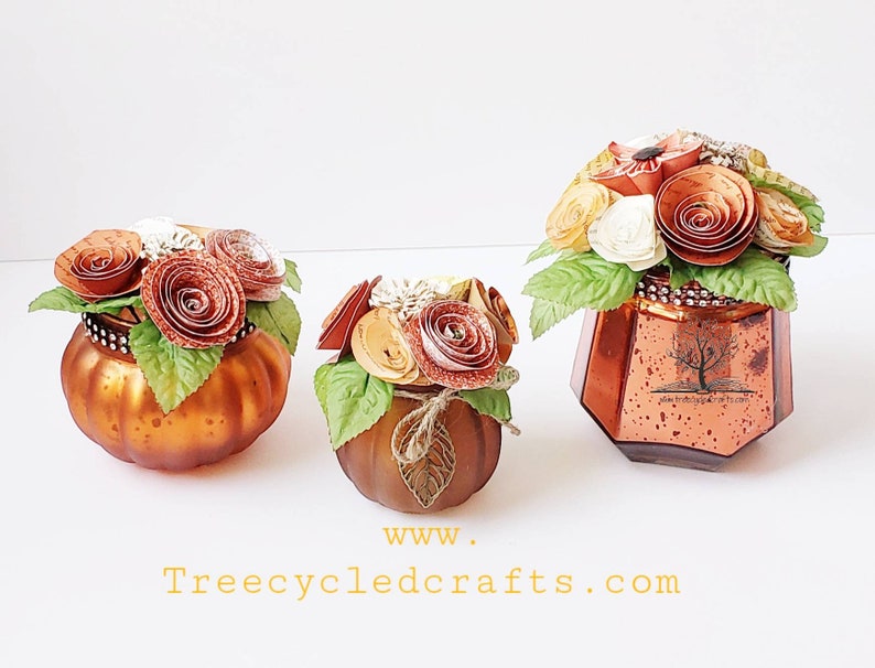 May include: Three glass pumpkins decorated with paper flowers in shades of orange, brown, and white. The pumpkins are all different sizes and have a rustic, vintage look. The largest pumpkin has a label that reads "Treecycled Crafts.com".