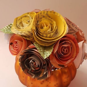 May include: A small orange pumpkin vase with a bouquet of paper flowers. The flowers are made from book pages and are in shades of yellow, orange, and brown. The vase is decorated with a glittery trim.