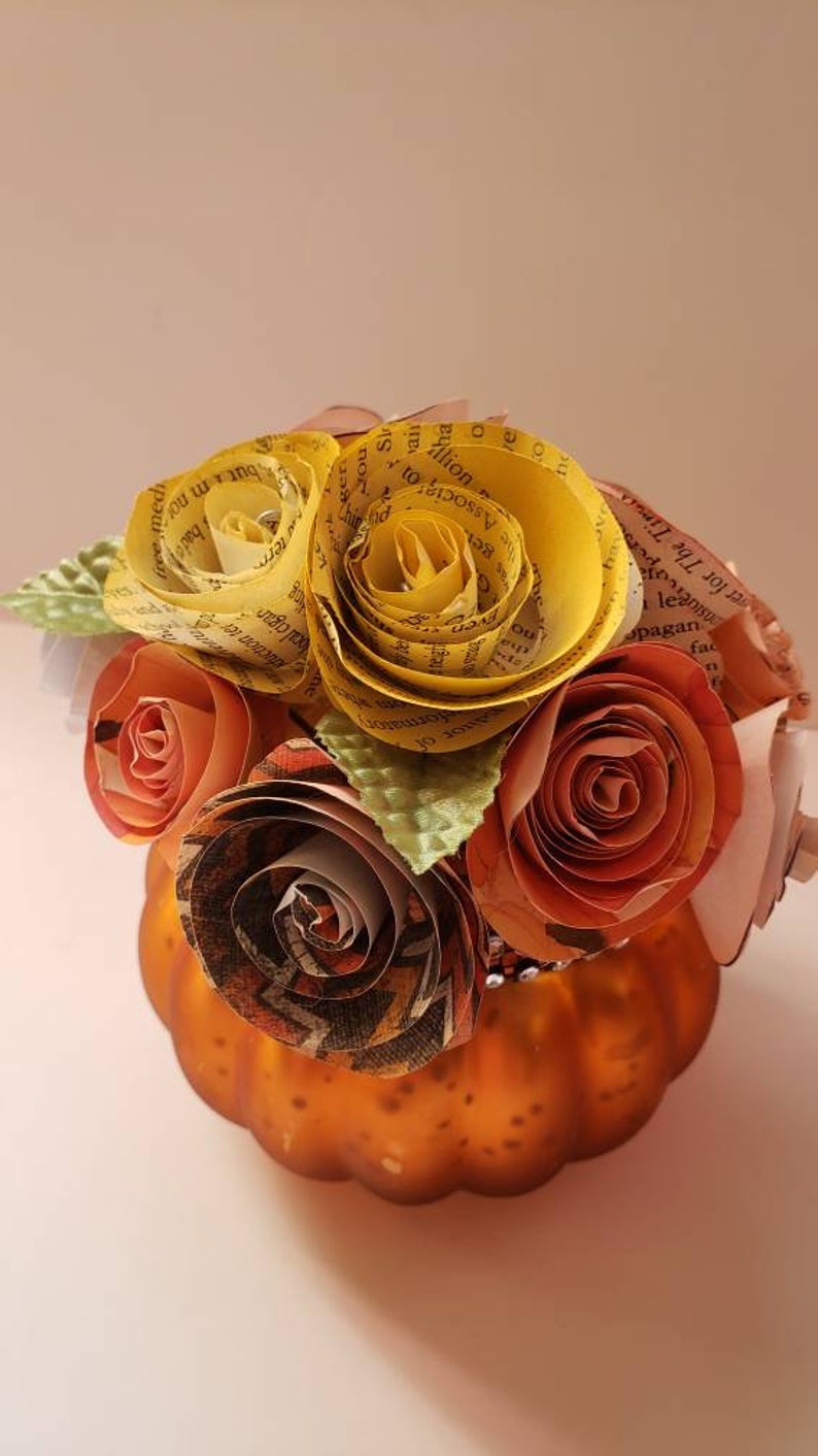 May include: A small orange pumpkin vase with a bouquet of paper flowers. The flowers are made from book pages and are in shades of yellow, orange, and brown. The vase is decorated with a glittery trim.