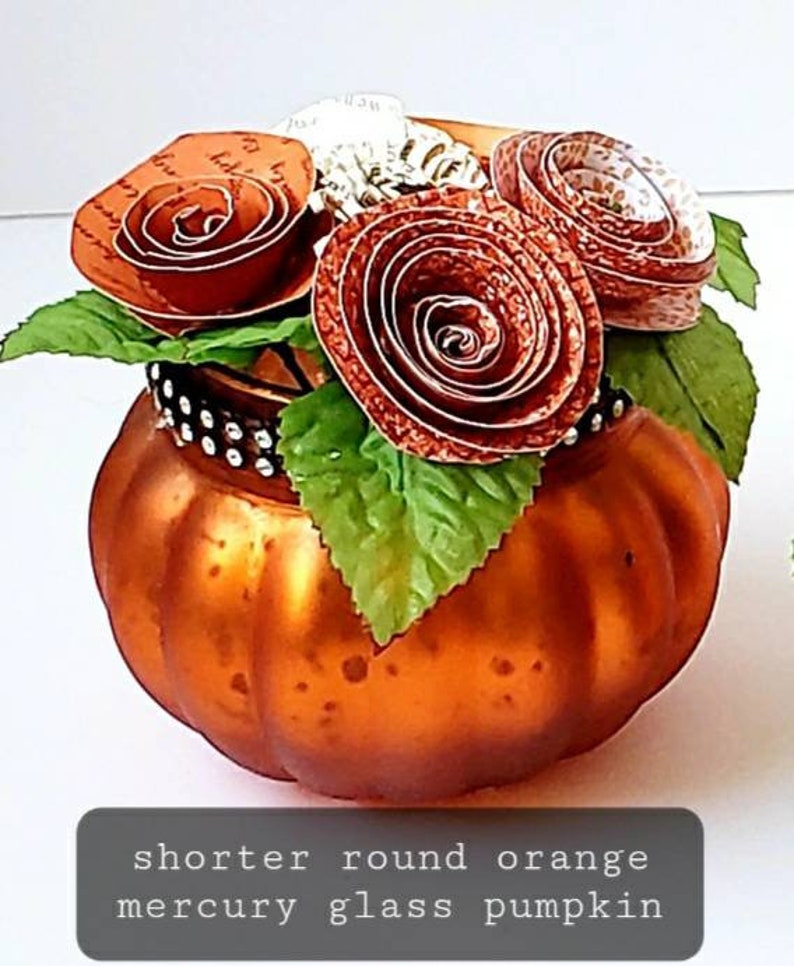 May include: A short, round, orange mercury glass pumpkin with a decorative arrangement of paper flowers. The flowers are made of brown paper and have a vintage look.