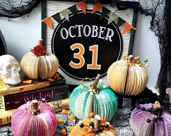 May include: A Halloween-themed display featuring several paper pumpkins in various colors, including pink, orange, and teal. The pumpkins are made from book pages and decorated with stems and embellishments. A sign reads "October 31."