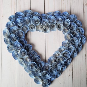 May include: A heart-shaped wreath made of blue paper roses. The roses are arranged in a gradient pattern, with the lightest blue roses at the center and the darkest blue roses at the edges.