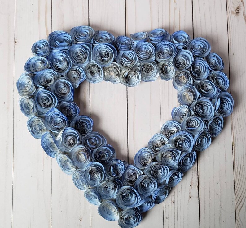 May include: A heart-shaped wreath made of blue paper roses. The roses are arranged in a gradient pattern, with the lightest blue roses at the center and the darkest blue roses at the edges.