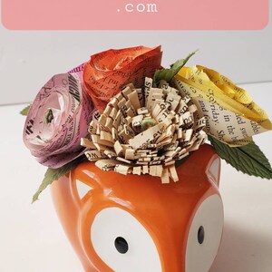 May include: A ceramic fox planter with a white glaze and black eyes. The planter is filled with paper flowers made from recycled book pages. The flowers are pink, orange, and yellow.