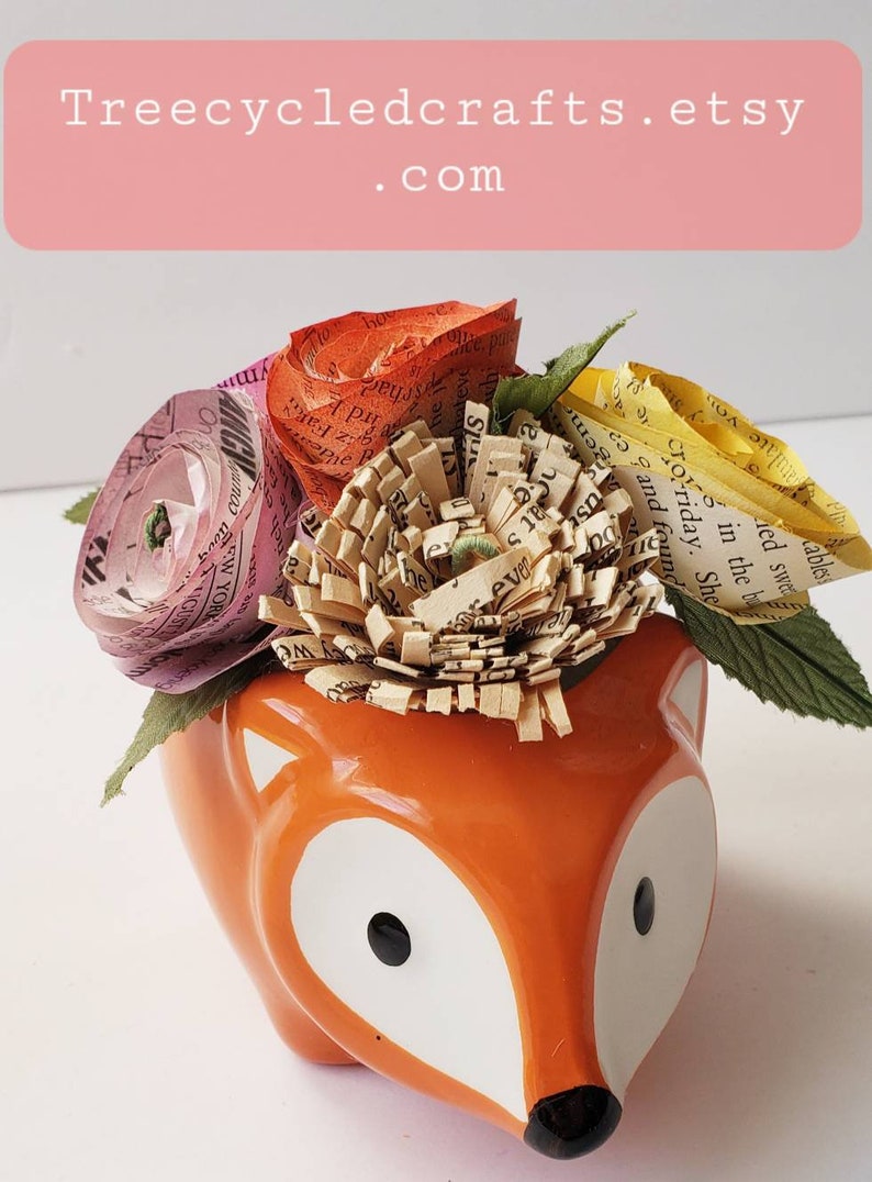 May include: A ceramic fox planter with a white glaze and black eyes. The planter is filled with paper flowers made from recycled book pages. The flowers are pink, orange, and yellow.