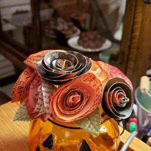May include: A glass pumpkin-shaped jar with a smiling face, decorated with a bouquet of paper flowers. The flowers are made from pages of a book and are in shades of orange, red, and black.