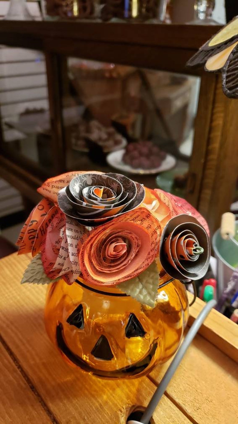 May include: A glass pumpkin-shaped jar with a smiling face, decorated with a bouquet of paper flowers. The flowers are made from pages of a book and are in shades of orange, red, and black.