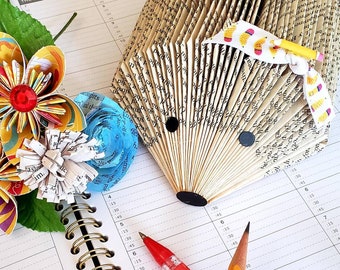 May include: A brown paper hedgehog sculpture made from folded book pages, with a white and yellow ribbon tied around its head. The sculpture is sitting on a white planner with a red pen and a yellow pencil.